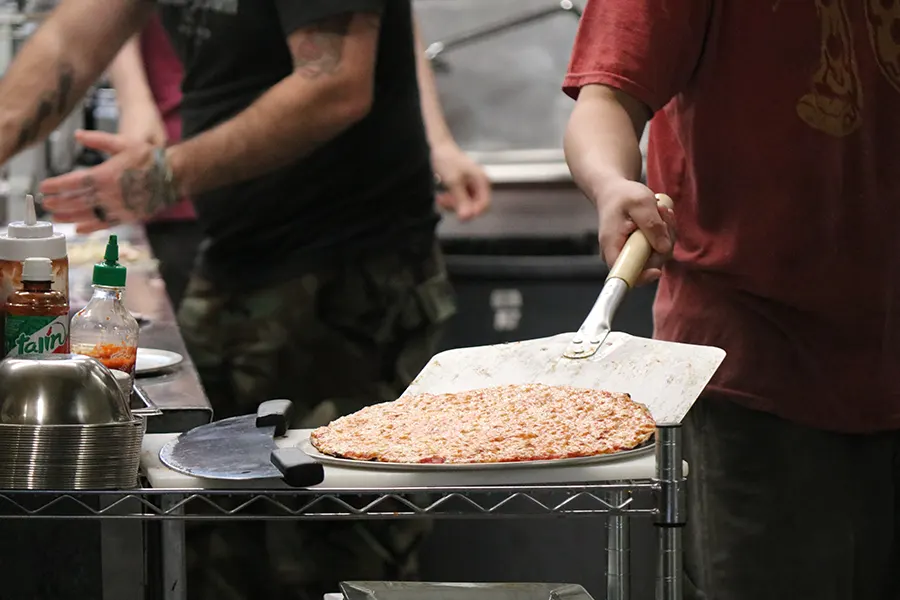 a photo back into the kitchen, where two chefs are hard at work. in the foreground, one cook scoops up a fresh-cooked and tasty-looking cheese pizza.