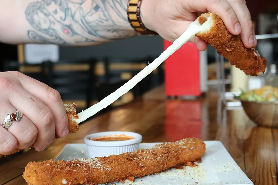 two hands pull a long strand of cheese from the inside of an enormous, crispy-looking mozzarella stick.